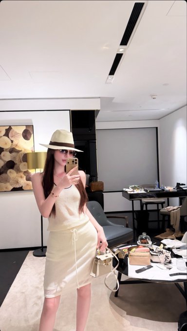 A woman wearing a hat, sunglasses, and a cream-colored dress takes a mirror selfie with her phone while holding a small handbag.