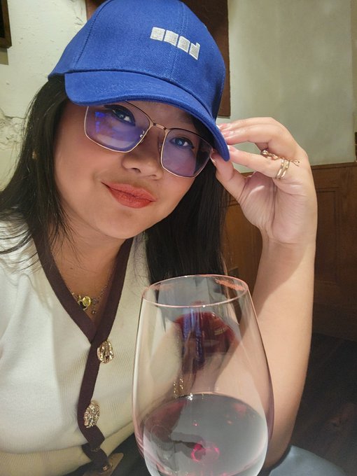 A selfie of a woman wearing a blue cap and gold-rimmed glasses with blue-tinted lenses, holding the brim of her hat and posing next to a wine glass in a restaurant setting.