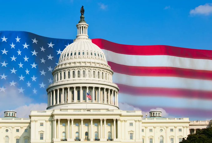 The U.S. Capitol building stands prominently with an American flag waving in the background against a bright blue sky.