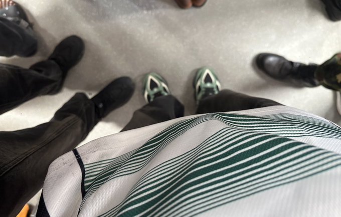 A first-person perspective looking down at someone's feet wearing green and white sneakers, surrounded by other people in dark shoes on a gray floor, with a white blanket or fabric with green stripes visible in the foreground.