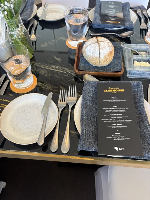A photo of a set dining table at 'BINANCE CLUBHOUSE BALI' featuring a detailed menu, fresh bread, plated dishes, and place settings on a dark marble surface.