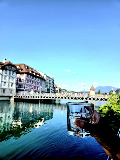 A hand raises a glass of water in a toast against a backdrop of Swiss architecture, a river, and distant mountains, visually embodying the post's message of trading financial losses for a life of clean water, scenic travel, and freedom.