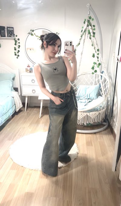 A woman takes a mirror selfie in a bedroom, wearing a gray spider-graphic tank top and baggy jeans, with a white hanging chair and ivy decor visible behind her.