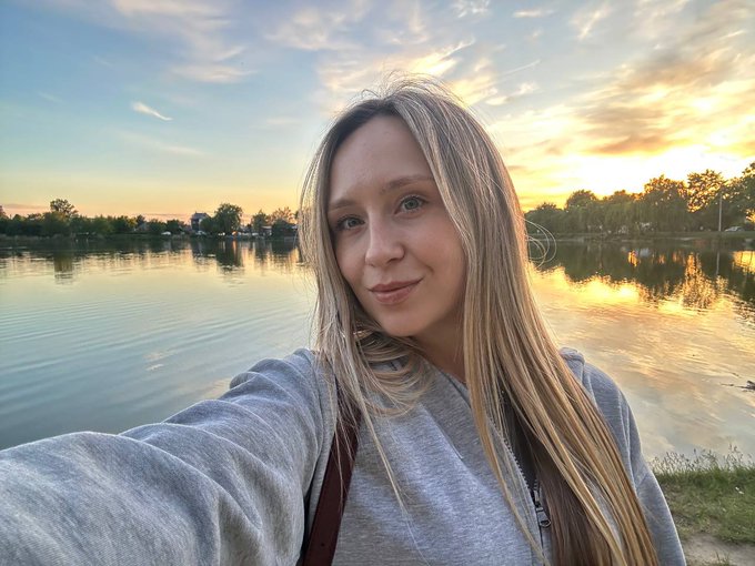 A blonde woman takes a selfie wearing a gray sweatshirt by a lake at sunset, with the orange sky reflecting on the water.