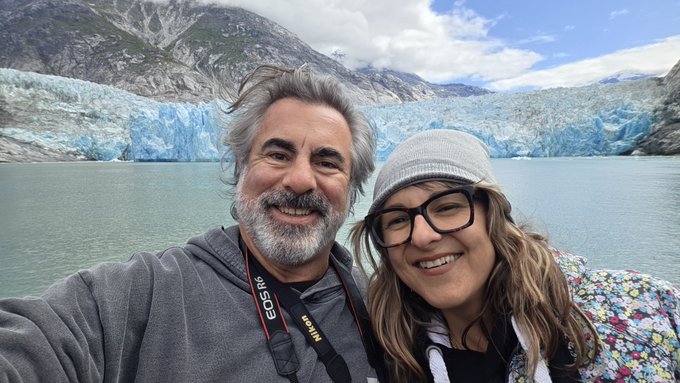 A selfie of two people, an older man with a gray beard and a younger woman with glasses, posing in front of a large blue glacier and mountains. A Nikon camera strap is visible.
