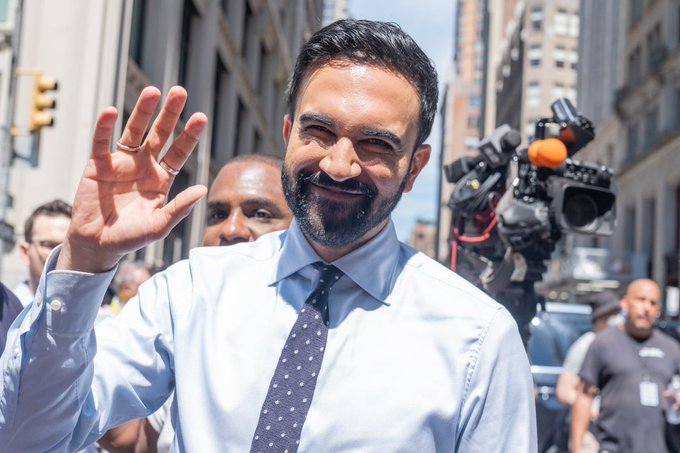 A man in a blue shirt and tie waving while walking down a city street, with a cameraman filming him.