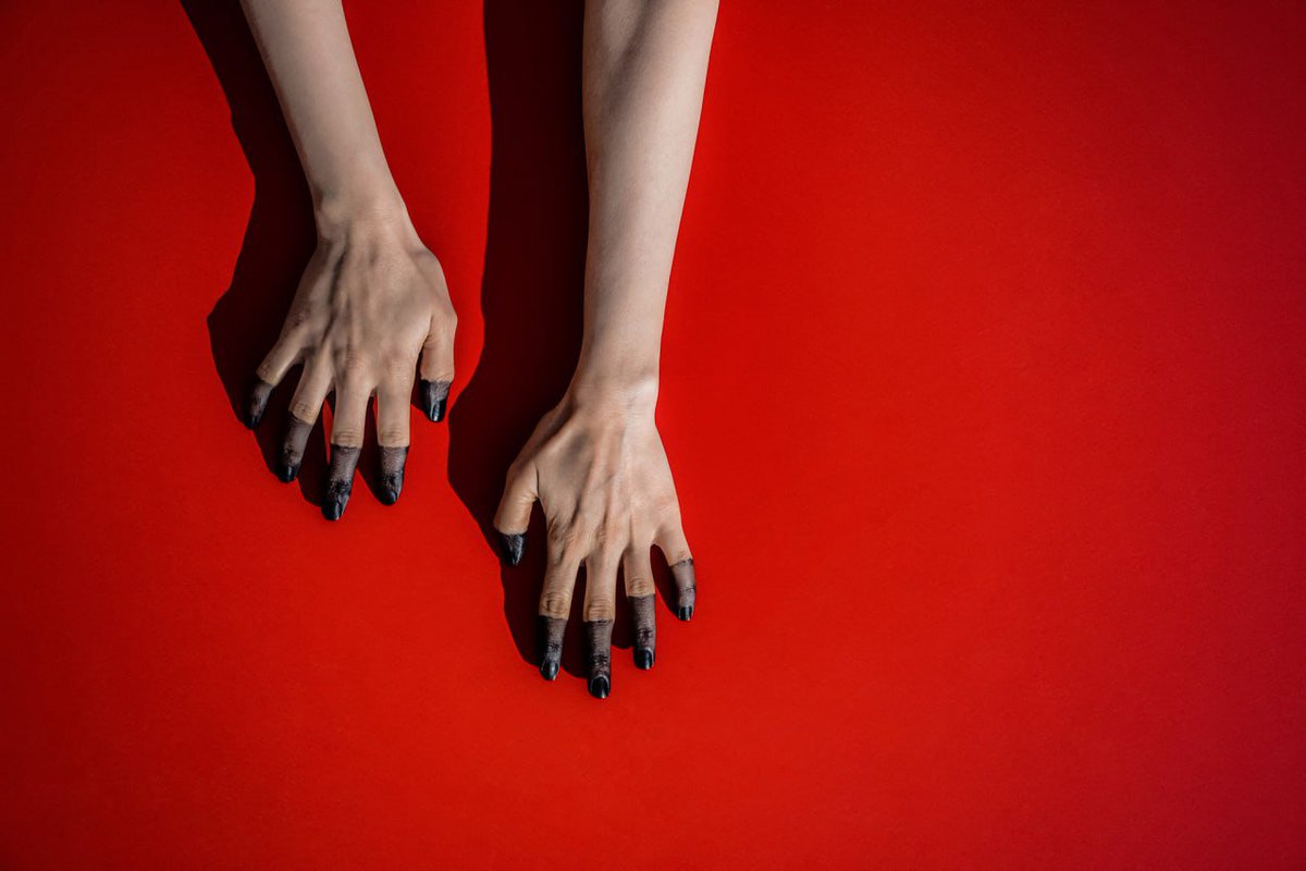 Two pale hands with black-stained fingertips hanging down against a vibrant red background, casting soft shadows.
