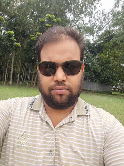 The image features a man wearing sunglasses, a shirt with white stripes, and a collar. He has a beard and appears to be smiling for the camera. The background of the photo shows trees, suggesting that he might be outdoors or near a park. The caption of the image reads "Good morning X legend! Learning to live anew in a serene morning. @NomismaNetwork Super bullish @useTria".