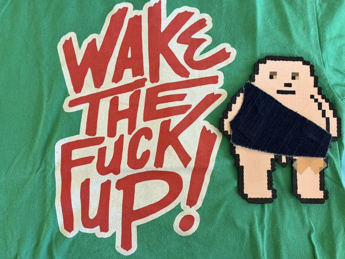 A graphic featuring the red text 'WAKE THE FUCK UP!' on a green fabric background, next to a pixel-art wooden figure of a person.