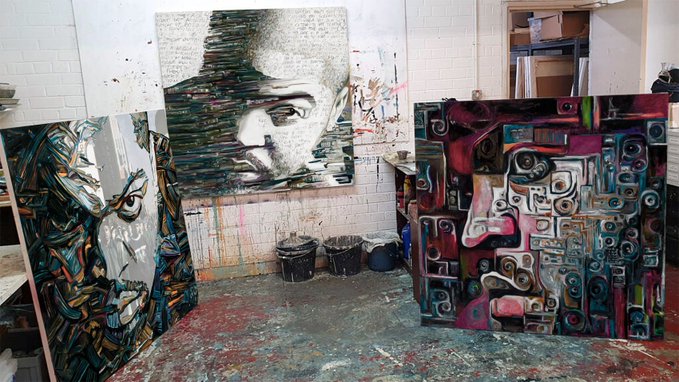 An artist's studio displaying several large canvas paintings featuring stylized portrait and abstract designs, with paint buckets and supplies visible on the floor.