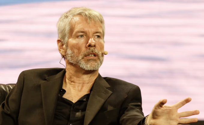A gray-haired, bearded man in a dark blazer speaks animatedly with a headset microphone, gesturing with his hand during what appears to be a public speaking event or interview, though the image itself shows no direct connection to the financial content about Strategy's $STRC yield.