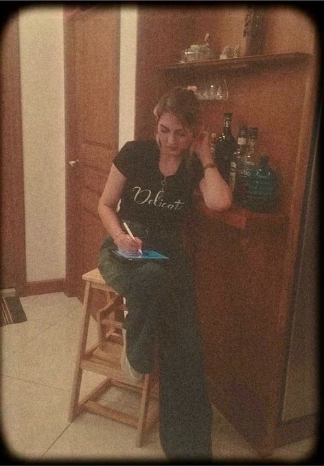 A vintage-filtered photo of a woman wearing a black 'Velo' t-shirt sitting on a wooden stool, writing in a notebook beside a wooden cabinet filled with liquor bottles.