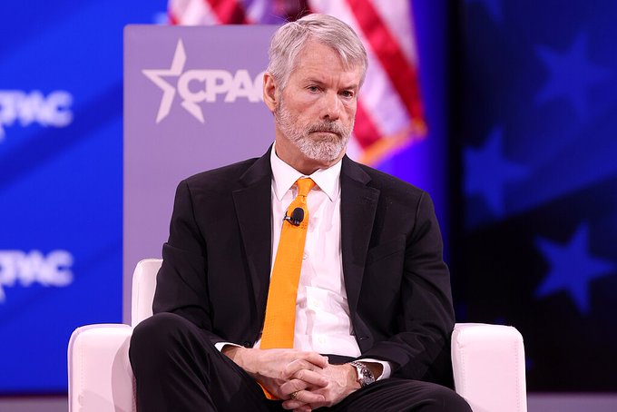 A gray-haired man in a black suit and bright orange tie sits with his hands clasped on stage at a CPAC event, with American flags and star-decorated backdrops visible behind him. The image appears to show Michael Saylor, the prominent Bitcoin advocate, lending visual support to his statement that Bitcoin has become the new hurdle rate on Wall Street.