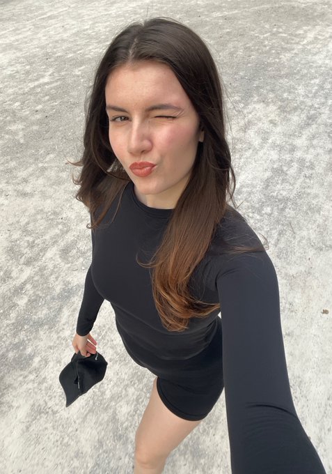 A young woman with long brown hair takes a selfie on a paved surface, winking and pursing her lips while wearing a black long-sleeved top.