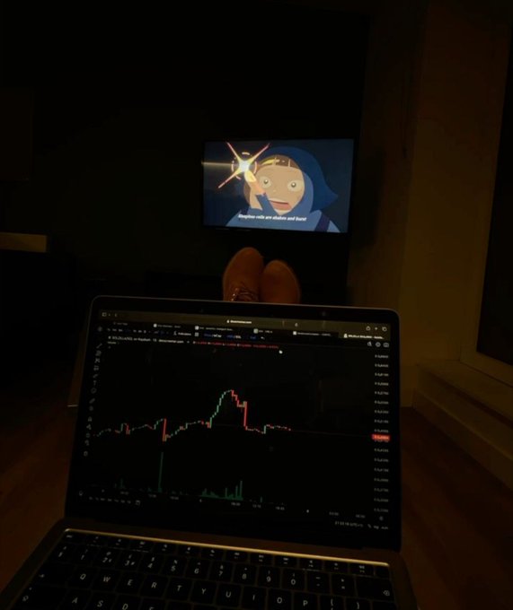 A person views a candlestick stock chart on a laptop in a dimly lit room while a TV plays an animated show in the background, with their feet propped up—capturing a relaxed morning routine of checking the markets.