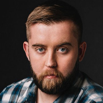 A portrait photograph of a young man with a beard, blue eyes, and short brown hair, wearing a blue plaid shirt against a dark background.