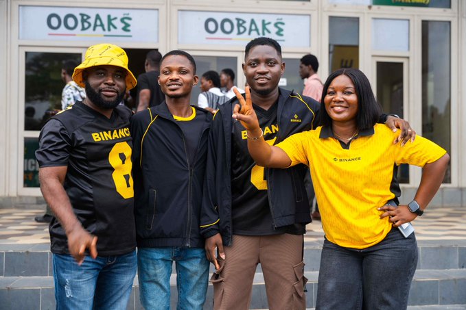 Four people pose together in front of storefronts with 'OOVAKE' signage; three wear black 'BINANCE' jerseys with one displaying 'BIMAN' and the number '8', while a woman wears a yellow Binance polo shirt.
