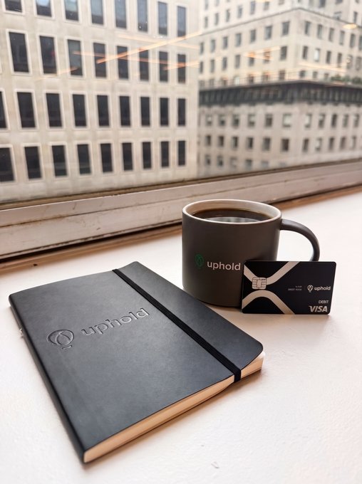 A cup of coffee sits next to an Uphold branded notebook and a Visa debit card, accompanied by a cityscape view. The items on the table relate to the post's mention of "Uphold" and "XRP rewards", likely referring to financial services.