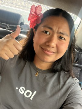 A woman with a red bow in her hair gives a thumbs-up while sitting in a car, wearing a gray t-shirt with partial text '.sol' visible.