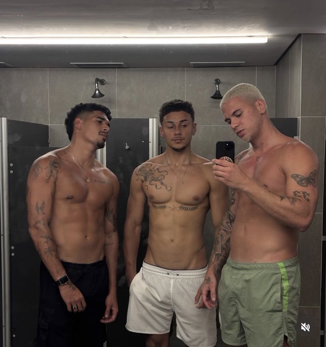 Three shirtless, tattooed young men pose for a mirror selfie in a locker room, with the man on the right holding a phone to capture the shot. The man in the middle wears white shorts and sports a chest tattoo, while the man on the left wears black shorts and has arm tattoos, and the blonde man on the right wears green athletic shorts.