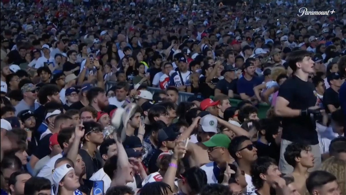 The image shows a large crowd of people gathered together, with some individuals holding up their phones or cameras. The crowd appears to be watching an event where Trump's crypto coin is being advertised during a coverage of the UFC fight.