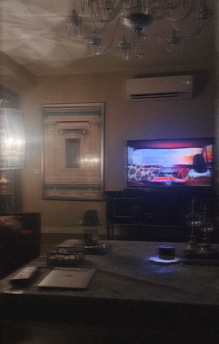 A dimly lit, elegant living room with a chandelier, an ornate mirror, and a TV showing a scene, capturing the cozy atmosphere of winding down during the final hour before The Oil Rigs mint. The relaxed, ambient setting reflects the post's mood of taking a moment to chill before a deadline.
