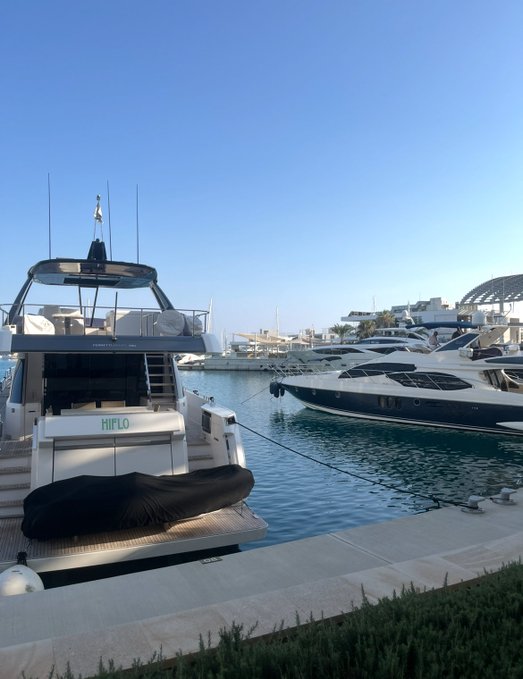 Sunny marina scene with a luxury yacht labeled 'HIFLO' docked in the foreground and other boats moored along the pier under a clear blue sky.