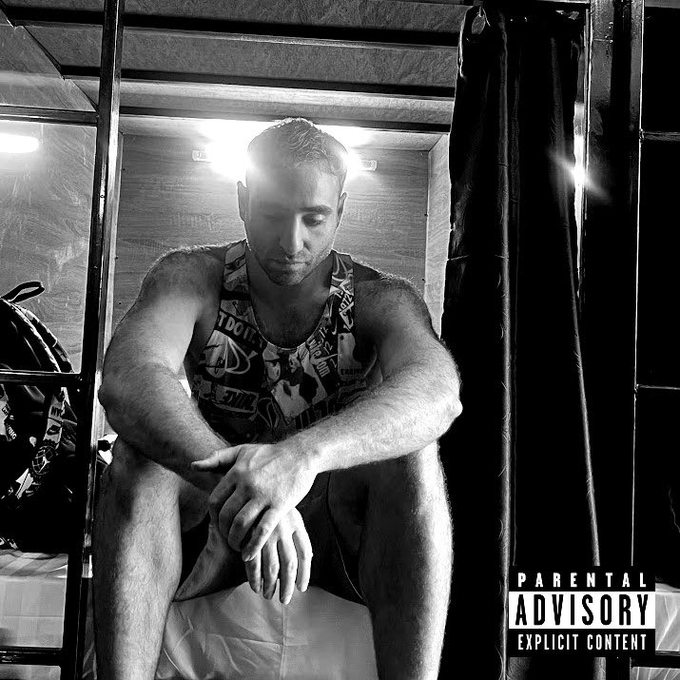 A black-and-white photograph of a muscular man sitting on the edge of a bunk bed in a dimly lit room, looking down at his hands with a 'Parental Advisory Explicit Content' label in the bottom right corner.