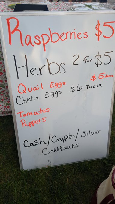 A handwritten whiteboard sign advertising farm stand prices: Raspberries $5, Herbs 2 for $5, Quail Eggs $5/dozen, Chicken Eggs $6/dozen, Tomatoes and Peppers, with payment accepted in Cash/Crypto/Silver/Goldbacks.