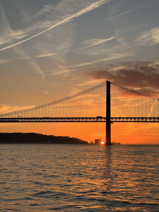 A scenic photograph of a large suspension bridge silhouetted against a vibrant orange sunset sky, with wispy clouds and the sun dipping near the horizon over a calm body of water.
