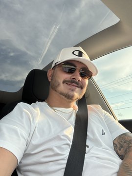 A man wearing sunglasses and a white Champion cap sitting in a car with a sunroof, wearing a seatbelt.