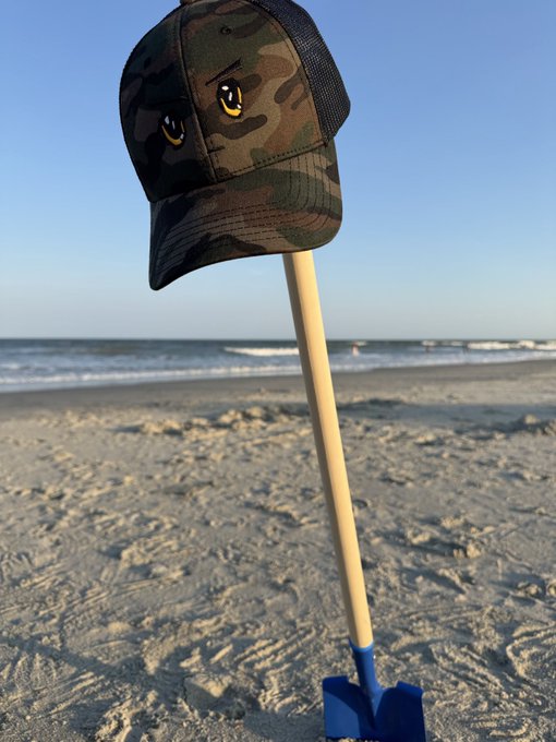 A camouflage cap featuring yellow eyes rests atop a blue shovel stuck in the sand at the beach, playfully illustrating the caption's theme of digging "down to Chinatown" with a "Griftevening" vibe.