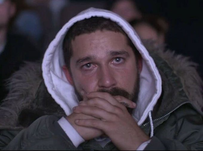 The image shows a man wearing a hooded jacket, sitting down with his hands clasped together over his mouth. He appears to be looking off into the distance or lost in thought. The background of the image features several other people who are also seated, but they do not seem to be the main focus of the scene. There is no text present in the image that directly relates to a post on social media.
