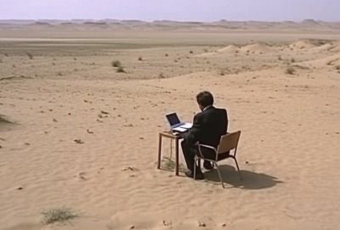 A businessman in a suit sits alone at a small table with a laptop in the middle of a vast, empty desert, illustrating the post's humor about tirelessly searching for a high-return investment even in the most barren conditions.