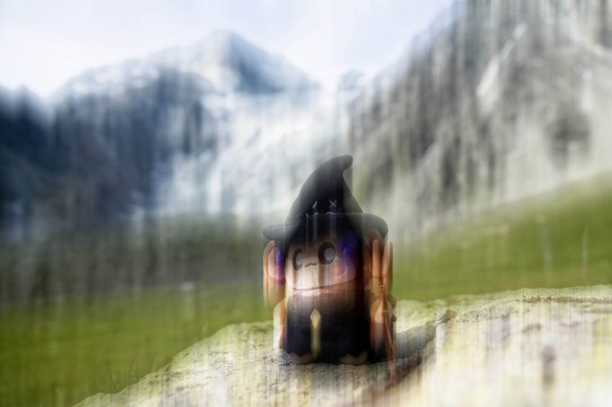 A small chibi-style wizard figure wearing a pointed hat sits on a rocky surface against a blurred background of snowy mountains and green grass.
