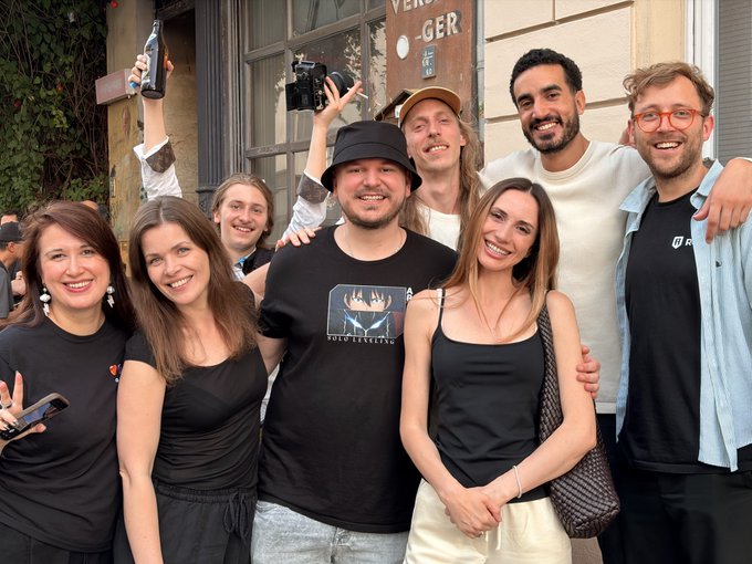 The image shows a group of people posing together, likely at an event or gathering. They are standing close to each other, smiling and enjoying themselves. The photo captures their camaraderie and happiness as they share this moment together. The background features a building with a window that has the word "GER" written on it, which could be related to the location of the event or simply an interesting detail in the scene.