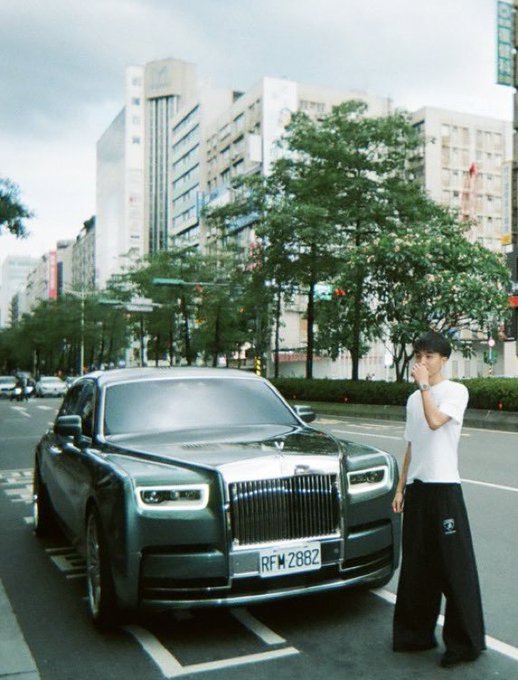 A photograph of a young man in a white t-shirt standing next to a black Rolls-Royce with license plate "RFM 2882" parked on a city street with buildings and trees in the background.