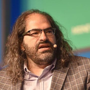 A man with long curly hair, glasses, and a headset microphone speaks at an event, appearing to be presenting. The image likely depicts David Schwartz in his role as CTO Emeritus at Ripple.