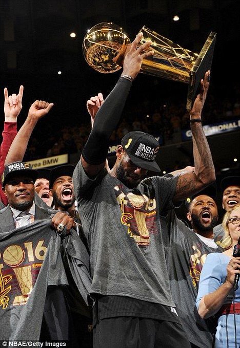 LeBron James holding up the NBA Championship trophy with his Cleveland Cavaliers teammates, wearing 'NBA Champions' hats and shirts commemorating the 2016 title.
