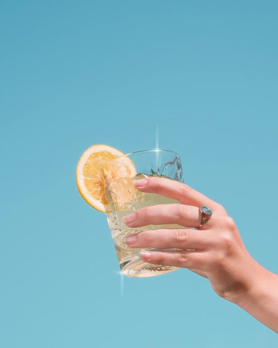 A hand holding a glass of yellow beverage with a lemon slice garnish against a bright blue background, with a subtle rainbow prism effect visible.