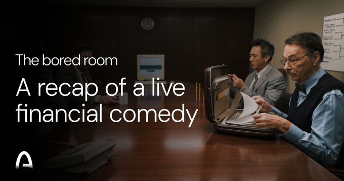 The image features two men sitting at a table with their laptops open, appearing bored as they work or browse the internet. The background shows an office setting with a clock hanging on the wall. The caption for this image reads "The Bored Room" and "A Recap of a Live Financial Comedy", suggesting that it is a humorous take on the concept of a bored room where people are working in finance-related roles, such as stockbrokers or financial analysts.