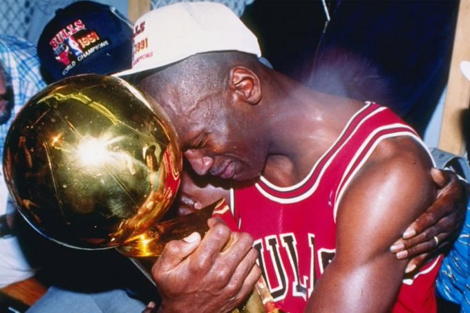 Michael Jordan, wearing a red Bulls jersey and a 'World Champions 1991' cap, emotionally embraces and kisses the championship trophy.