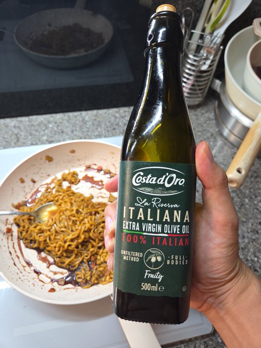 A hand holds a 500ml bottle of "Ostad'Oro La Riserva Italiana" Extra Virgin Olive Oil in front of a bowl of noodles with a dark sauce, on a kitchen counter.
