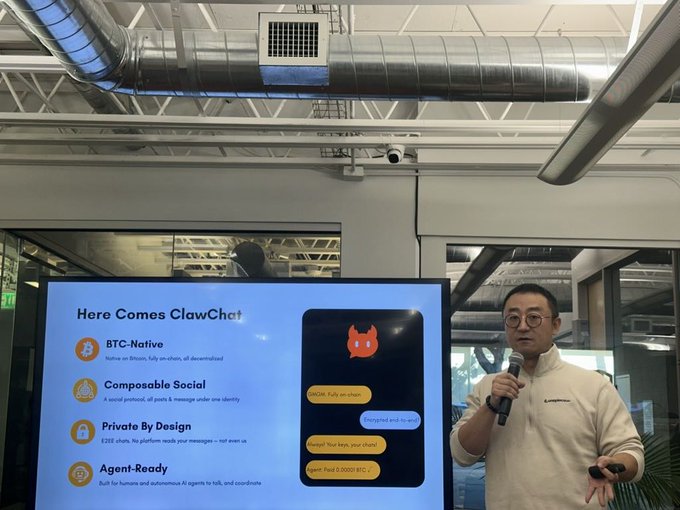 The image features a man standing next to a large screen displaying information about a Bitcoin programmable platform called " clawshat". The man appears to be giving a presentation or speech, possibly at an event related to Bitcoin technology. The background of the image shows a white ceiling with pipes running along it, suggesting that this might be taking place in a building with a ventilation system.