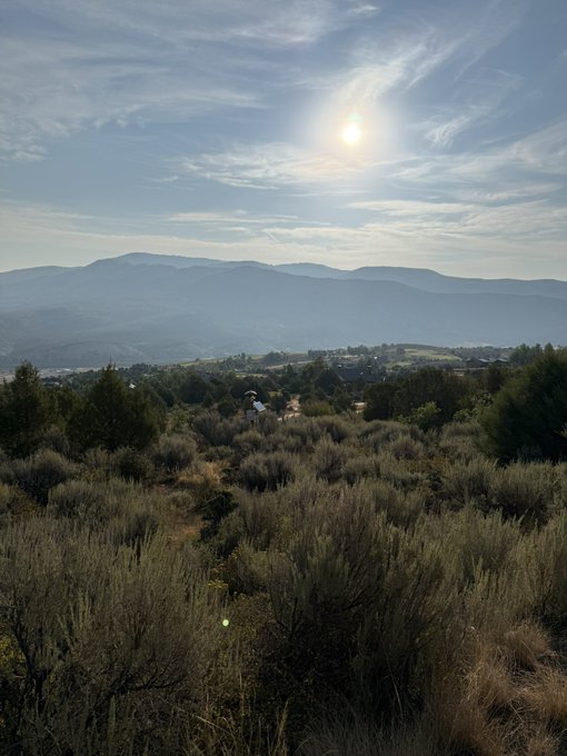 A landscape photograph showing a hilly area with sagebrush in the foreground, mountains in the background, and a bright sun shining in a partly cloudy sky.