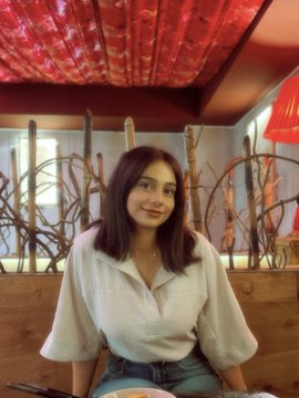 A young woman with dark hair wearing a white collared blouse smiles softly at the camera in what appears to be a restaurant booth with red ceiling decorations.