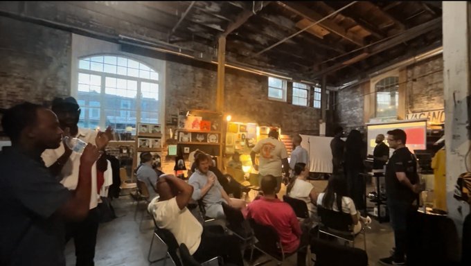 A group of people gathered in a industrial-style loft space with exposed brick walls, large windows, and clothing displays, possibly at a fashion event or showroom.