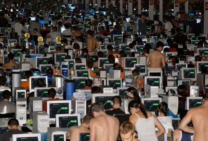 A large, crowded internet café or gaming center filled with rows of desktop computers, where many shirtless young men are sitting and standing while using the machines.