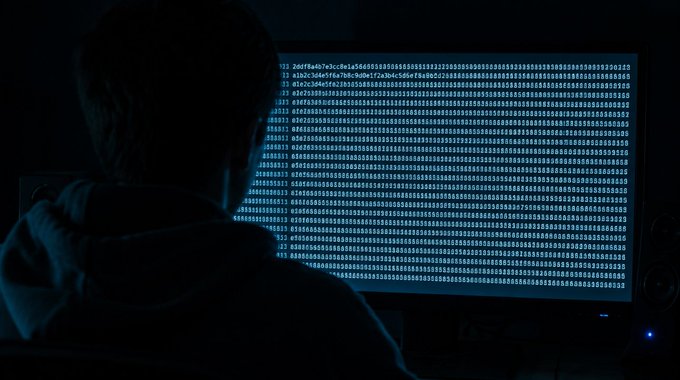A silhouette of a person in a dark hooded sweatshirt sitting in front of a monitor filled with scrolling lines of hexadecimal code in blue.