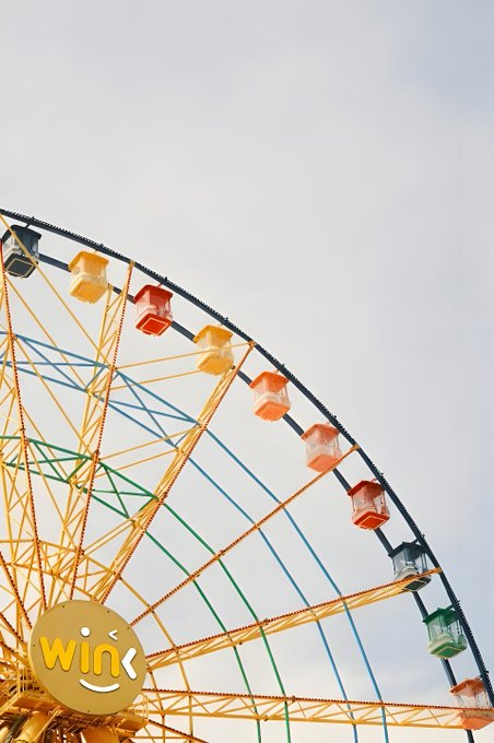 A vibrant Ferris wheel with multicolored gondolas stands against a pale sky, featuring a smiley face logo with the text "Wink" in the lower left. The whimsical ride visually contrasts with the post's discussion of automating solar-powered radio station grants on TRON, symbolizing the joyful community impact that transparent broadcasting infrastructure can deliver.